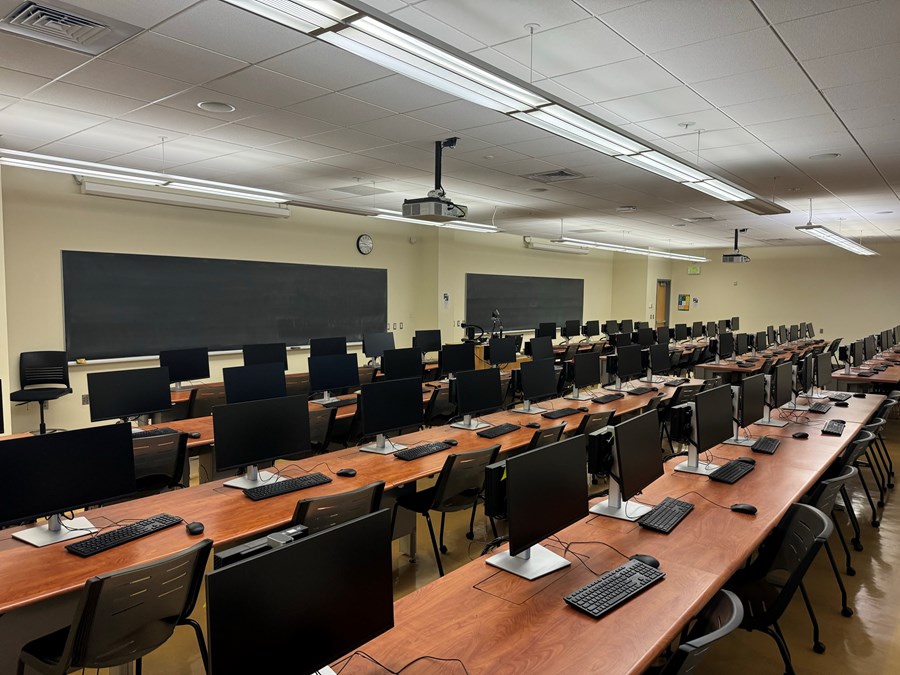 Image of Chiles 225 from the back of computers on desks with rolling chairs. Chalkboards and projector screens are against the far wall