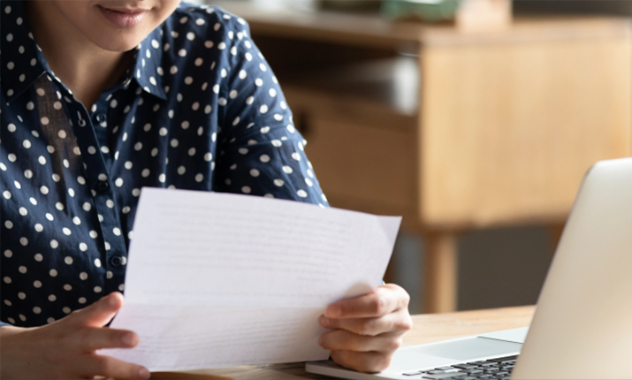 A person in a blue shirt with white dots is examining a piece of paper, laptop computer to the right.