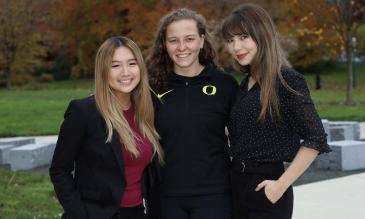 Three students standing close together, smiling at the camera, in front of a lawn and trees in the background